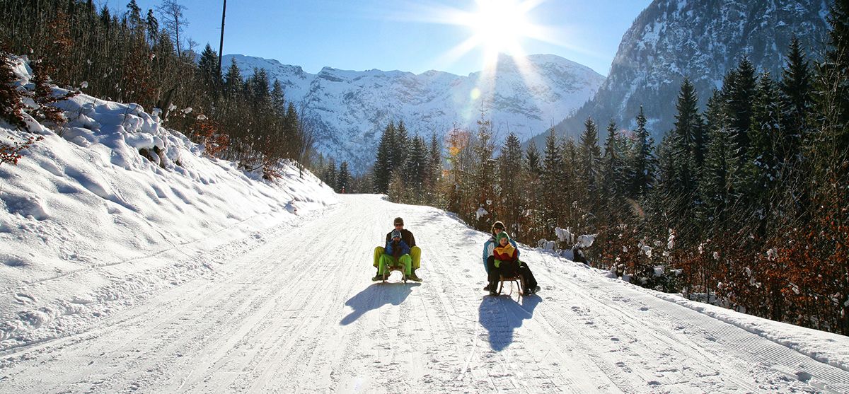 A snowy mountain road with two people sitting on sleds.