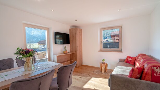 Una sala de estar moderna con una mesa de comedor de madera, sillas grises, un sofá con cojines rojos, un televisor montado en la pared y ventanas amplias con vistas a la montaña.