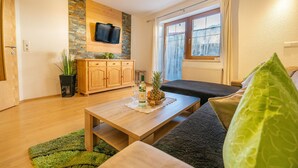 Una sala de estar acogedora con una mesa de centro de madera, un sofá, un televisor montado en una pared de piedra y una ventana con cortinas.