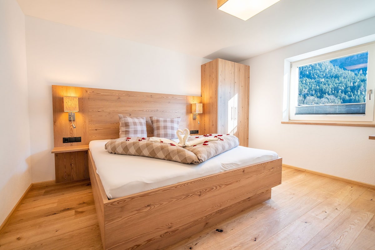 A bedroom with a wooden bed, two bedside lamps, a window with a forest view, and a painting on the wall.