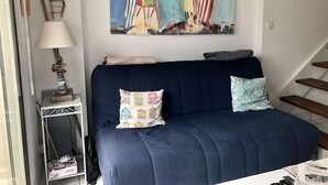 Ein Wohnzimmer mit einem dunkelblauen Sofa, zwei Kissen, einer Lampe und einem Gemälde mit bunten Häusern.