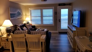 A living room with a television, a painting, a dining area with chairs, and a view of the ocean through the door. - Welcome to "Ocean Sunrise". Renovated direct ocean front private end unit.  (Wildwood Crest)