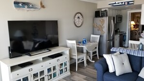 A living room with a flat-screen TV, a dining table with chairs, a sofa, a kitchen area with a refrigerator, and a wall clock. - Welcome to "Ocean Sunrise". Renovated direct ocean front private end unit.  (Wildwood Crest)