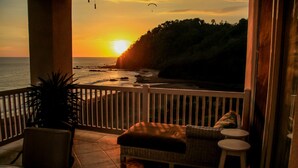 A balcony with a view of the sunset over the ocean, a wicker sofa, and a small table.
