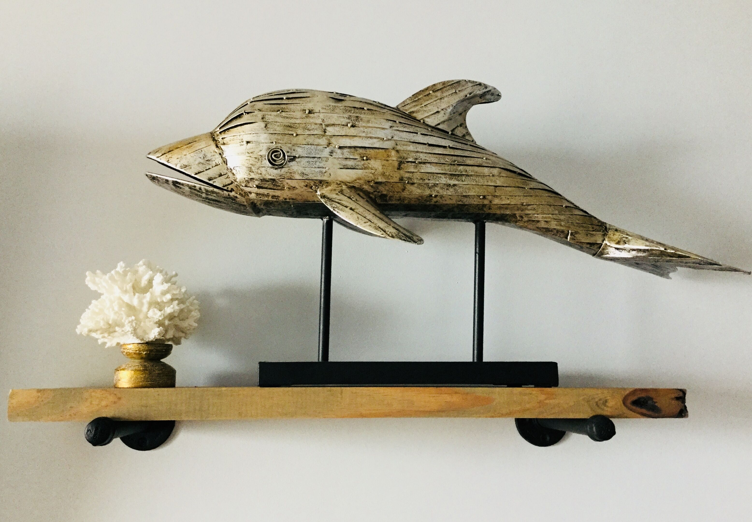 Une sculpture de poisson en bois, posée sur un socle noir, avec un petit vase blanc sur une étagère en bois située en dessous.