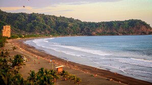A beach with a curved shoreline, palm trees, and a building in the background.