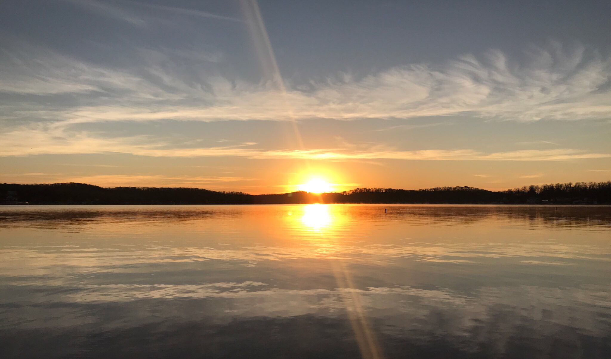 Ein ruhiger See bei Sonnenuntergang, die Sonne spiegelt sich im Wasser, umgeben von einem bewaldeten Ufer.