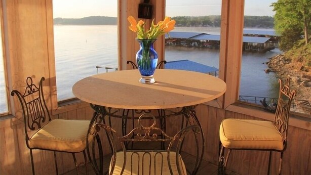 A wooden cabin with a round table and two chairs, a vase with flowers, and a view of a lake and dock.