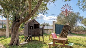 Ein Spielhäuschen aus Holz, eine Schaukel, ein Picknicktisch und ein Windrad auf einer grünen Wiese.