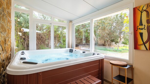 Ein Whirlpool in einem sonnendurchfluteten Raum mit großen Fenstern, einer hölzernen Sitzbank und einem kleinen Hocker.