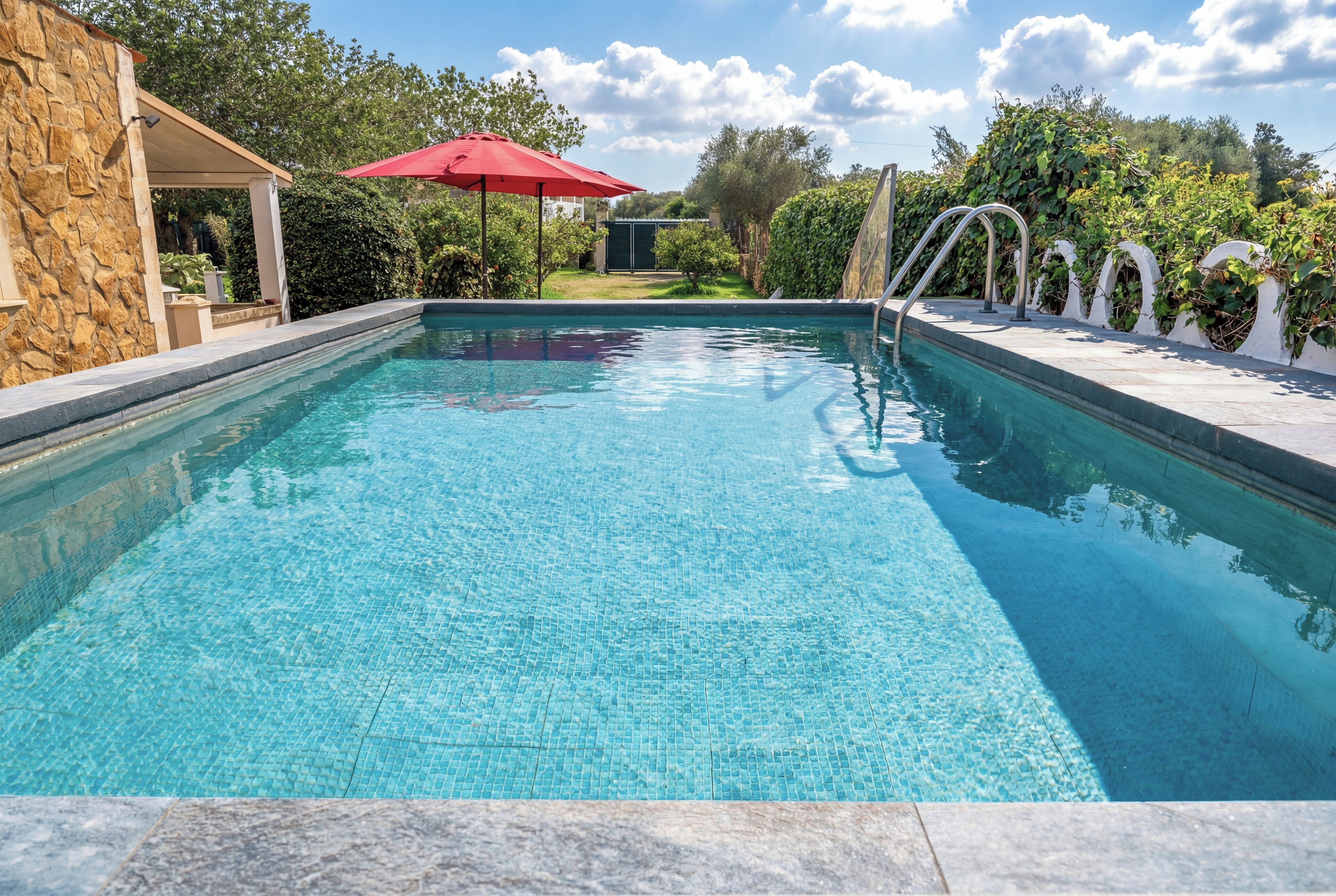 A clear blue swimming pool with a stone wall on one side and a covered patio area with a red umbrella.