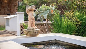 Eine Steinstatue eines Kindes, das Wasser trinkt, umgeben von einem kleinen Teich mit Seerosen, inmitten eines Gartens mit verschiedenen Pflanzen und einem Gebäude im Hintergrund.