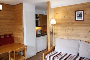 Een gezellige kamer met een houten wand, een bed met gestreept beddengoed, een houten tafeltje en een kitchenette op de achtergrond.