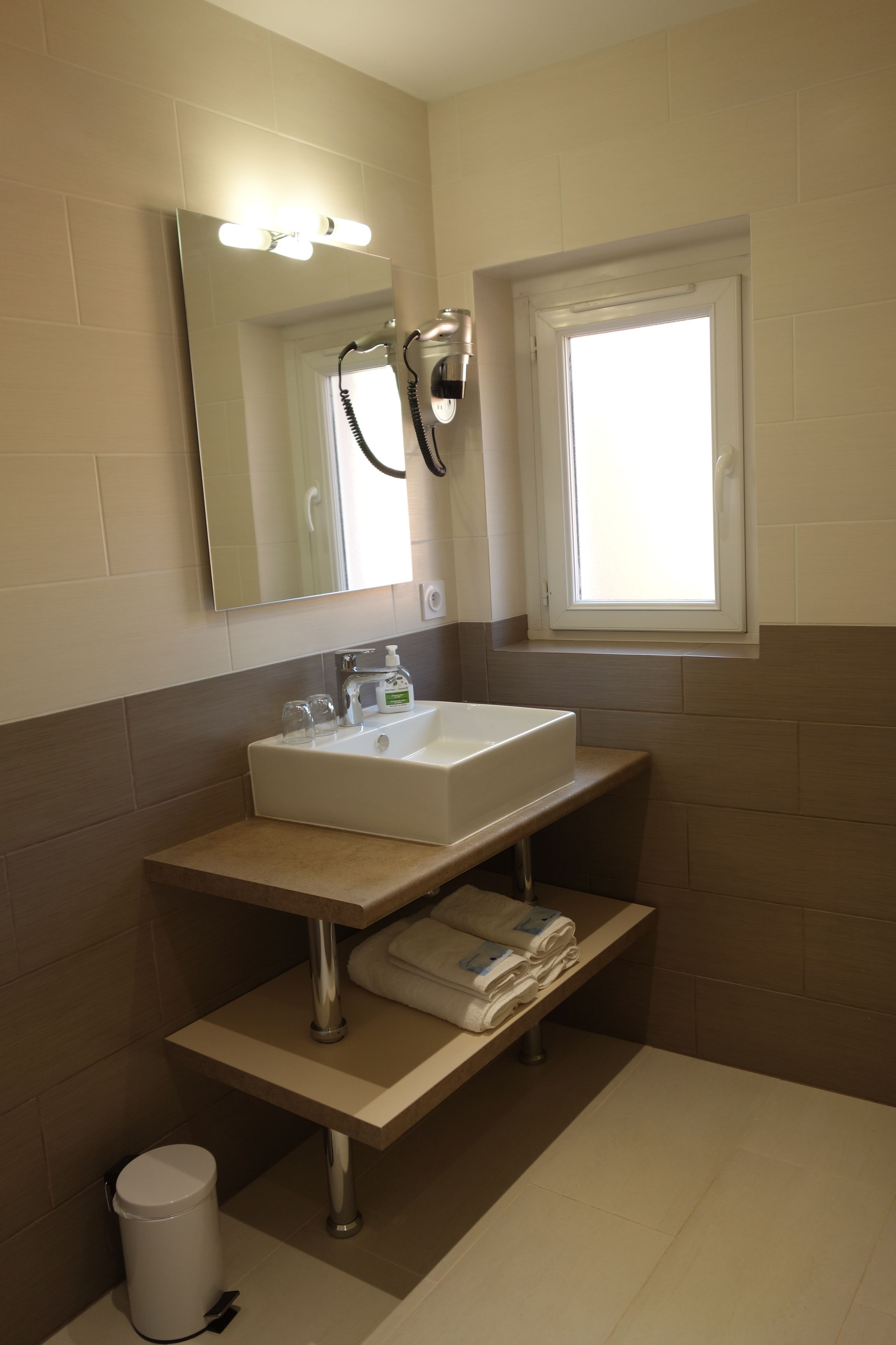 A bathroom with a white sink, a mirror with lights, a window, and a trash can.