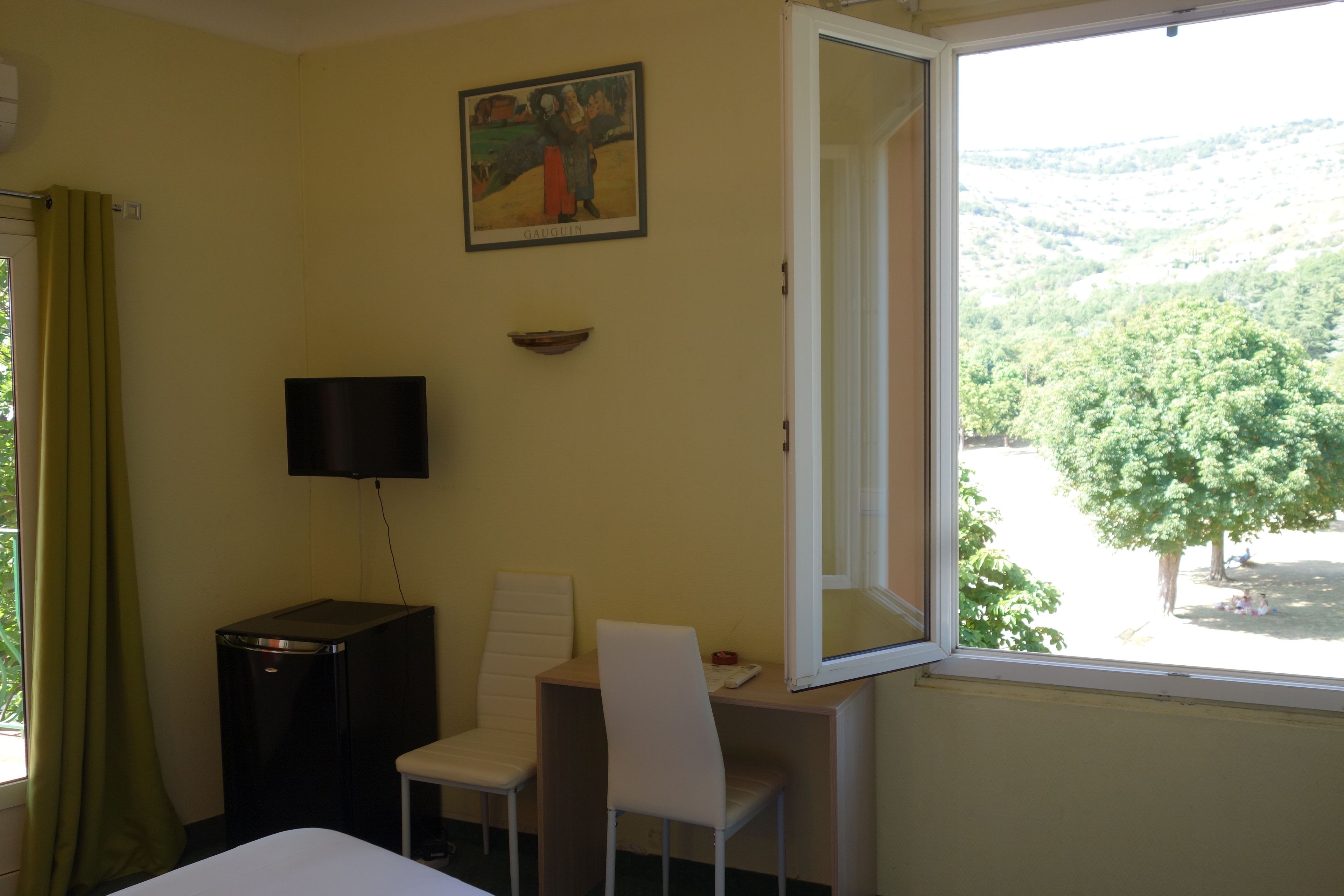Habitación de hotel con cama, escritorio con silla, televisión, un cuadro en la pared y ventana con vista a árboles y colinas.
