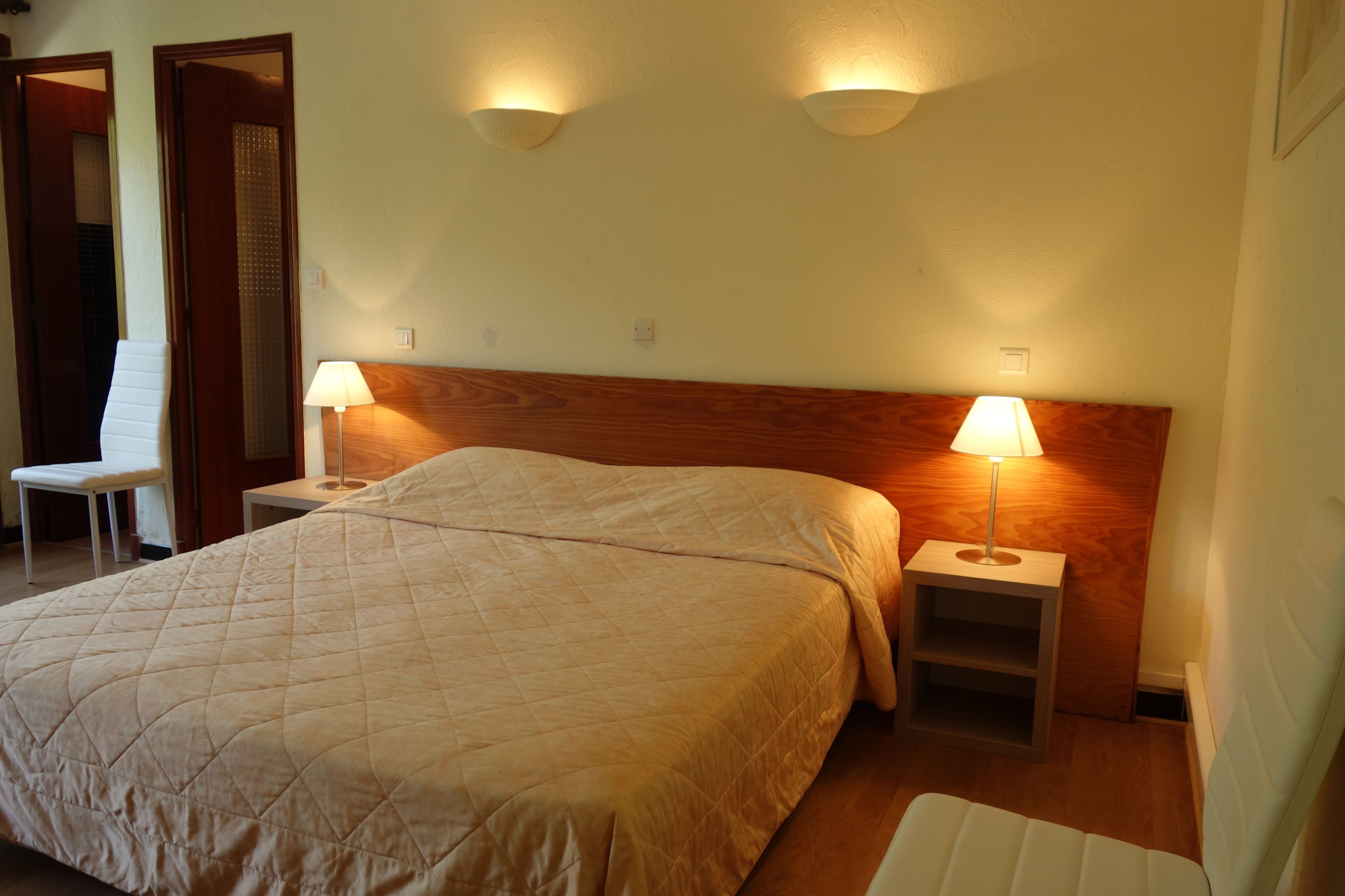 A bedroom with a large bed, two bedside lamps, a wooden headboard, and a white chair.