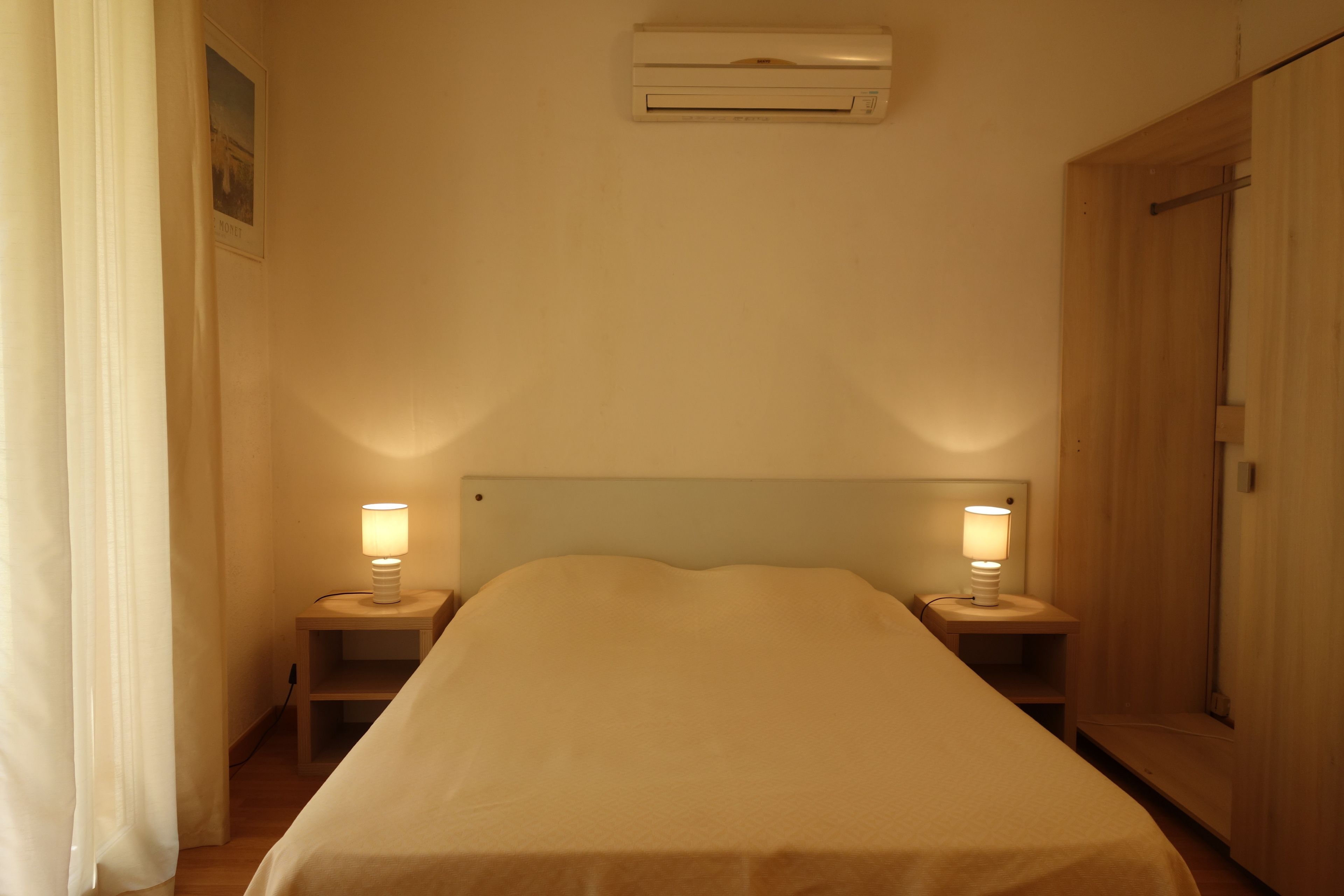 A single bed with a headboard, two bedside lamps, an air conditioning unit above the bed, and a wooden wardrobe to the right.