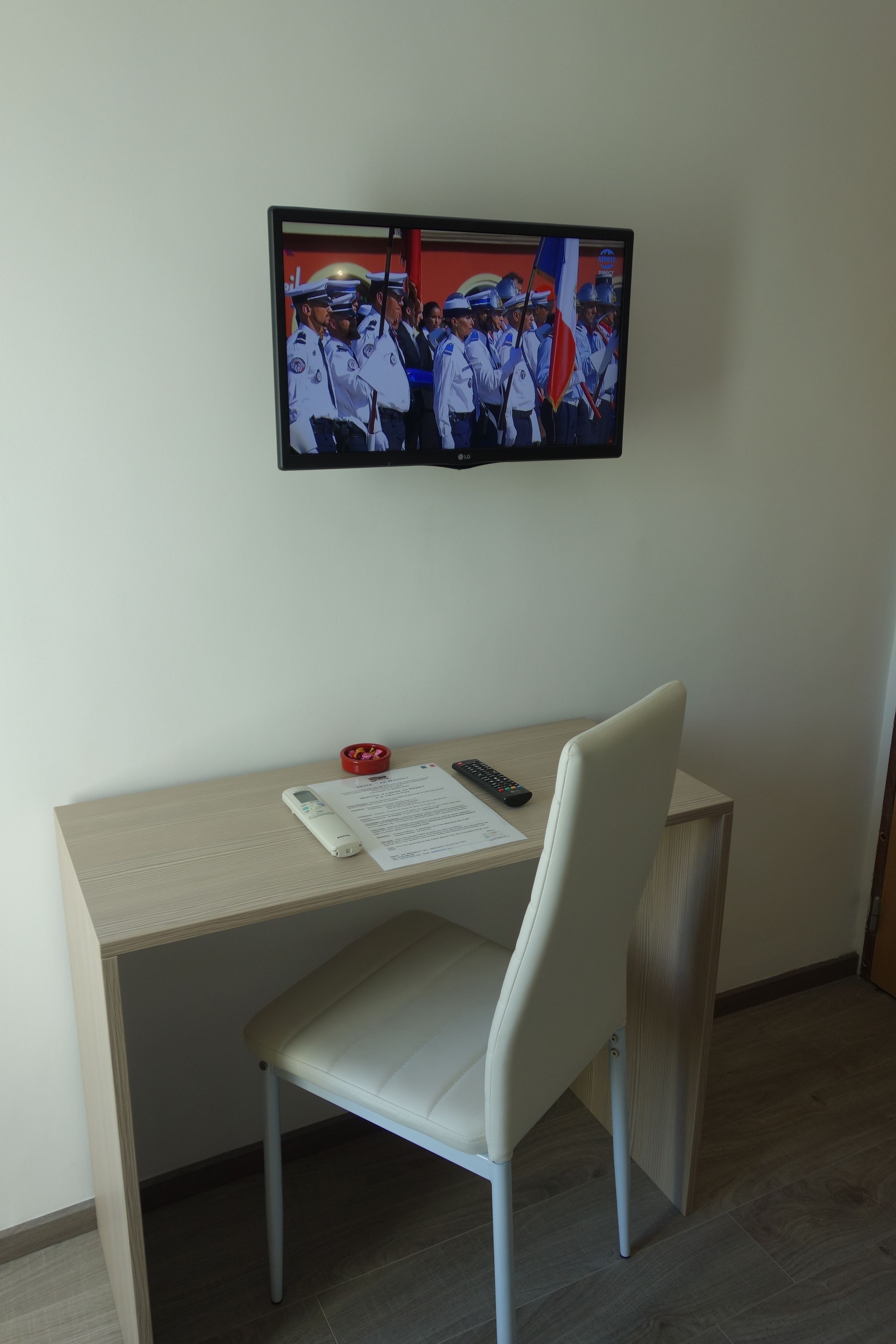 Een kamer met een bureau, stoel en een televisie aan de muur, waarop een groep mensen in uniform te zien is.