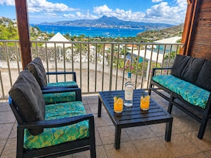 Uma varanda com vista para o mar, duas cadeiras, uma mesa e dois copos de sumo de laranja.