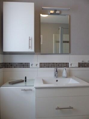 Une salle de bain avec un lavabo blanc, un miroir et un meuble. Il y a deux prises électriques sur le mur et une bouteille sur le lavabo.