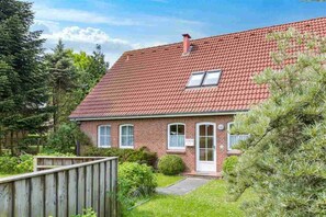 Une maison à deux étages, construite en briques, avec un toit recouvert de tuiles rouges, un petit porche à l’avant et une clôture en bois.