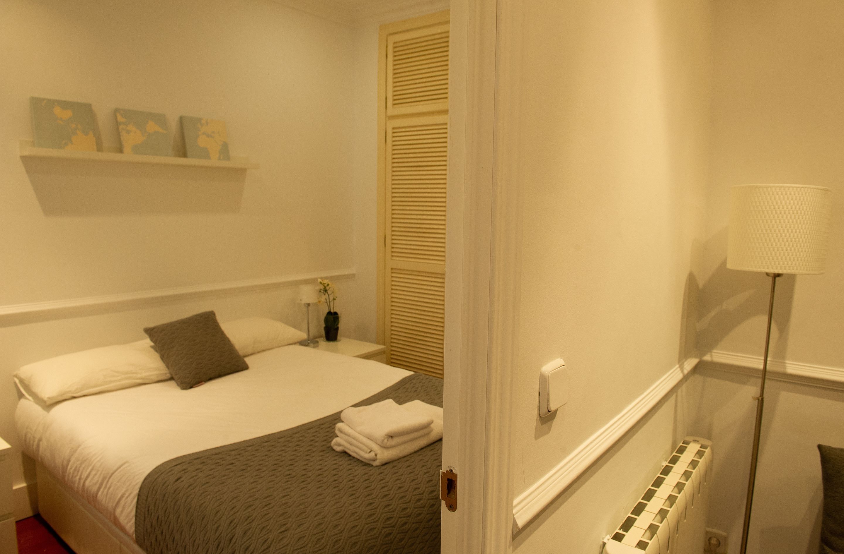 Una cama bien tendida con una manta gris y almohadas, una mesita de noche con una lámpara, y un estante en la pared con objetos decorativos.