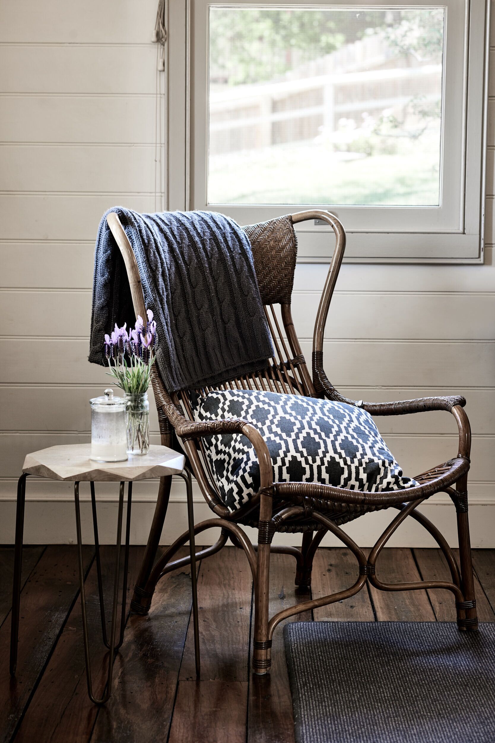 Una silla de mimbre con un cojín estampado, una mesita con un florero y vistas a una terraza de madera a través de una ventana.
