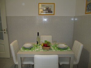 Une table à manger recouverte d’une nappe à motifs floraux verts, avec des chaises blanches et un panier de fruits.