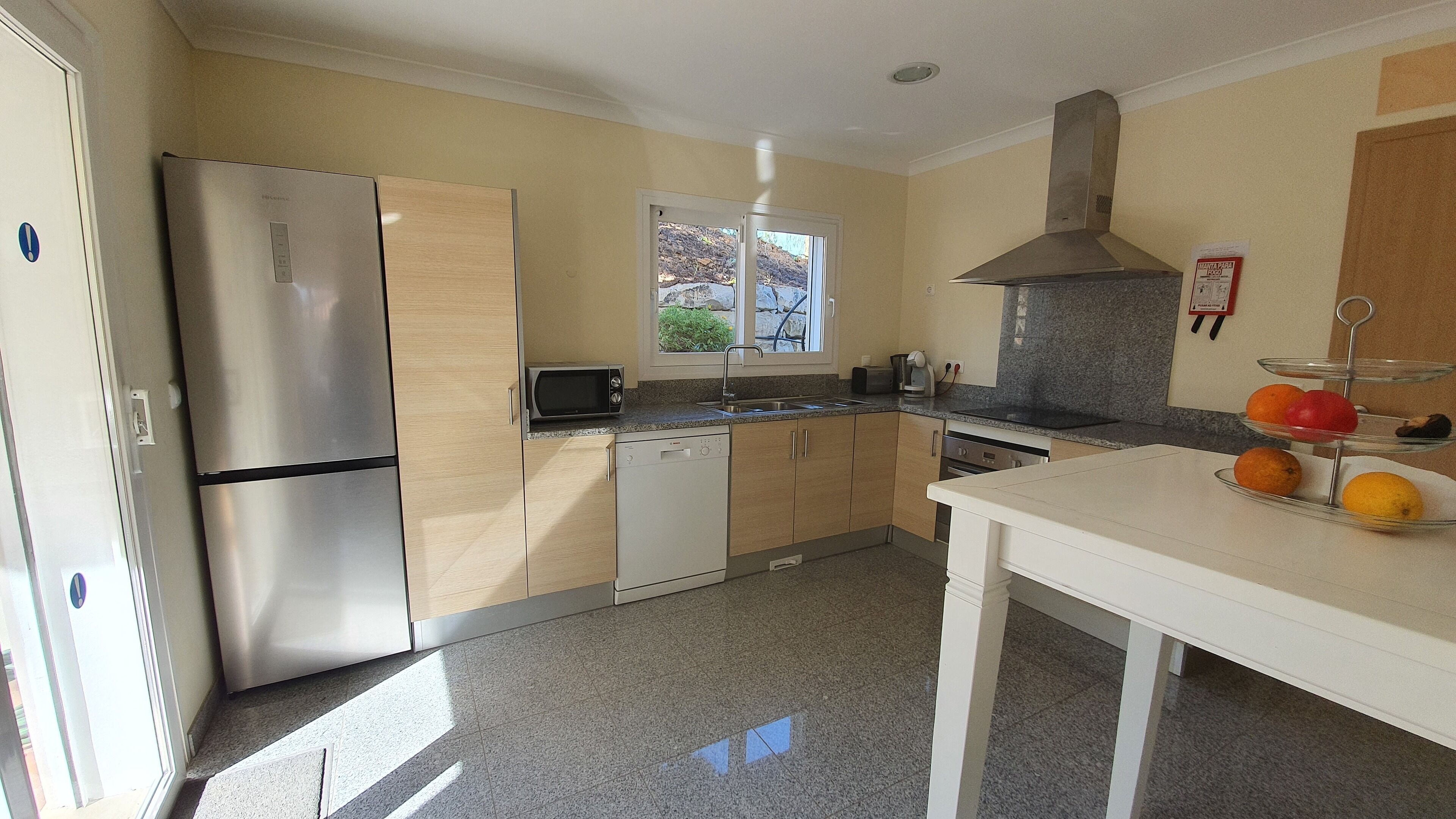 A compact kitchen with a stainless steel refrigerator, a dishwasher, and a sink. There's a window above the sink, and a three-tiered fruit stand on the counter.