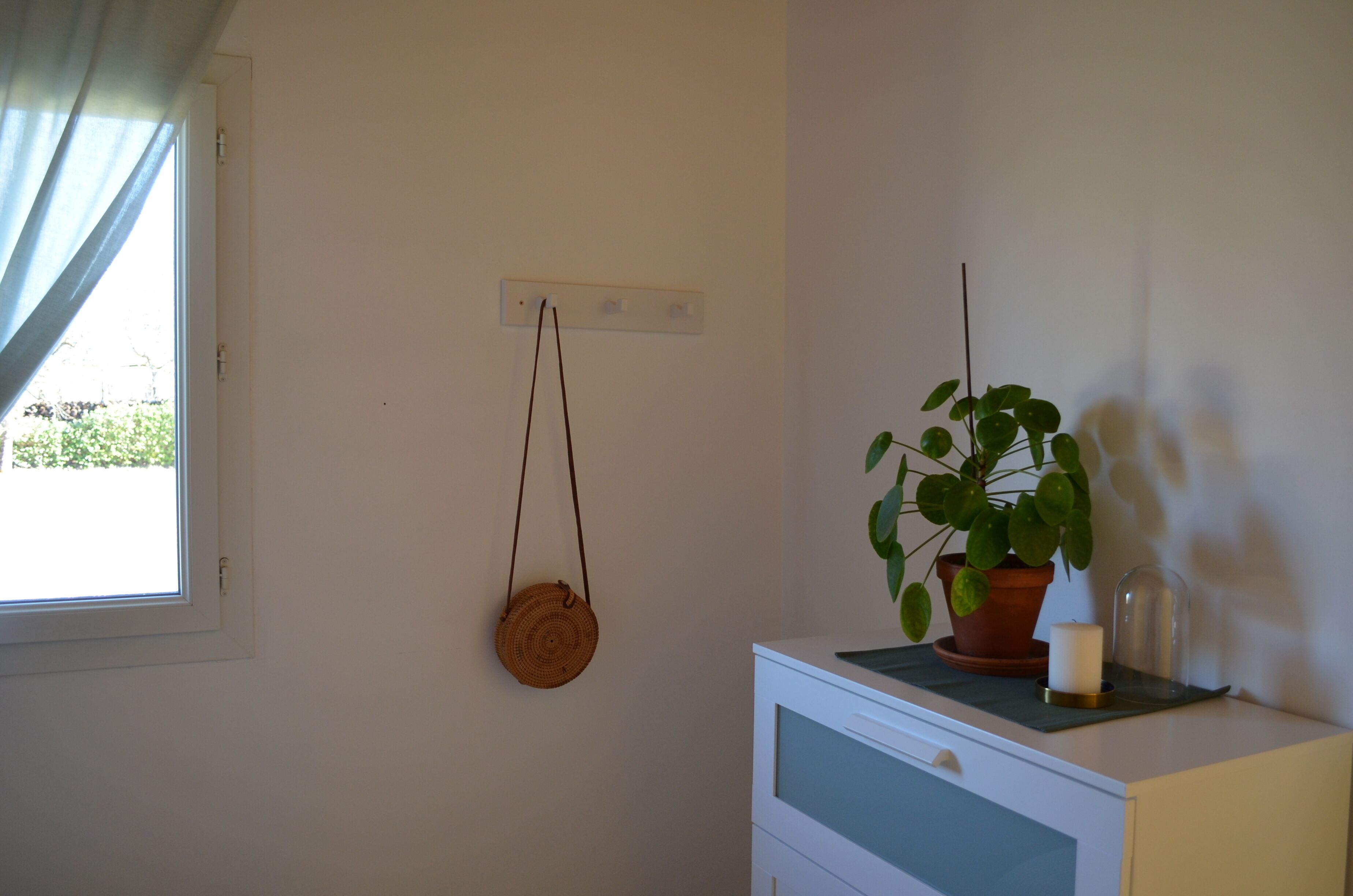 Ein Zimmer mit Fenster, einem Wandlichtschalter, einem kleinen Topfpflanzen, einer runden gewebten Tasche und einer weißen Kommode.