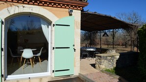 An open door to a room with a dining table and chairs, a stone wall, and a covered outdoor area with a grill. - Sunny holiday in the quiet countryside with pool. (Lalbenque)