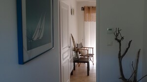 Un pasillo con un cuadro de un velero, una silla, un jarrón con ramas y un termostato en la pared.