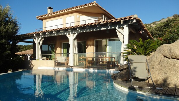 Un área junto a la piscina con una piscina cristalina, un porche cubierto con mobiliario exterior y una gran roca.