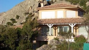 Un edificio de dos pisos con techo de tejas, rodeado de árboles y terreno rocoso.