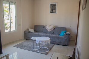 A living room with a grey sofa, a small white table with a lace tablecloth, and a blue pillow.
