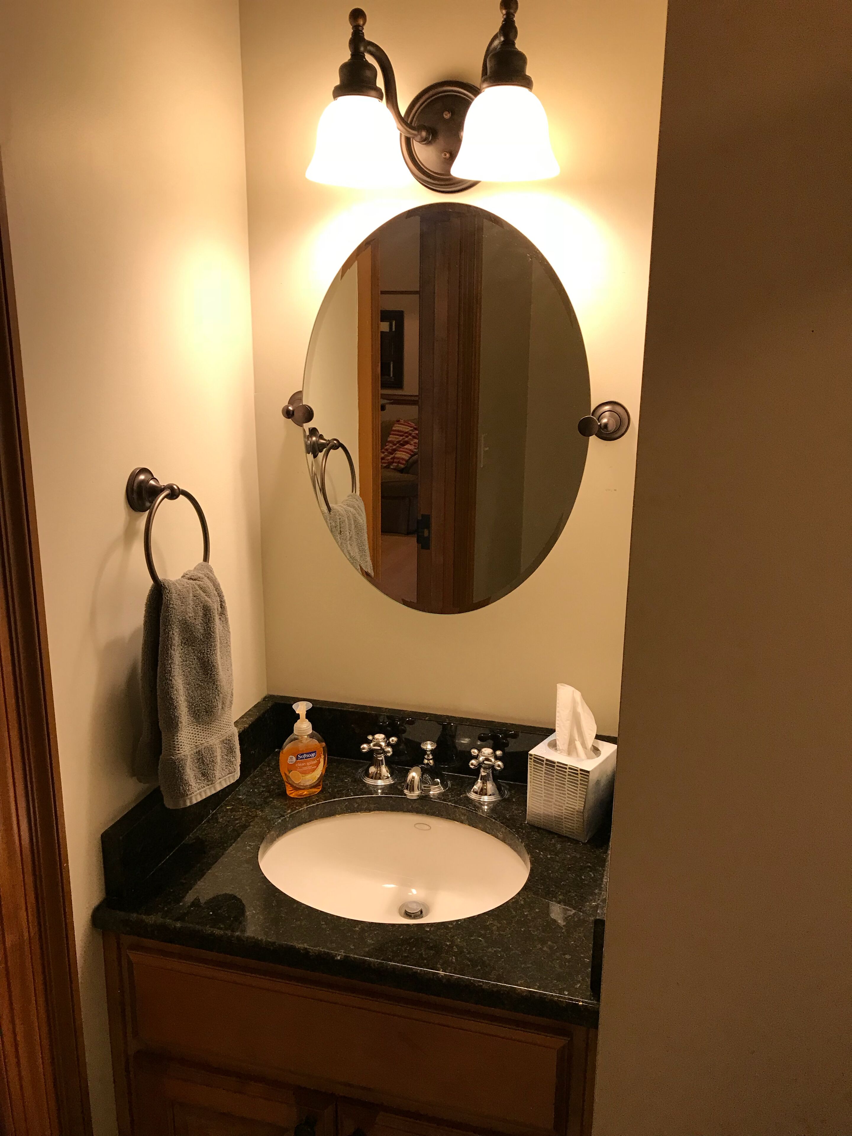 Un baño con un espejo redondo, dos luces empotradas en la pared, un perchero, un lavabo con dispensador de jabón y una caja de pañuelos.
