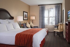 Une chambre d’hôtel avec un grand lit, deux lampes de chevet, un bureau avec une chaise, une télévision et une fenêtre avec des rideaux à motifs.