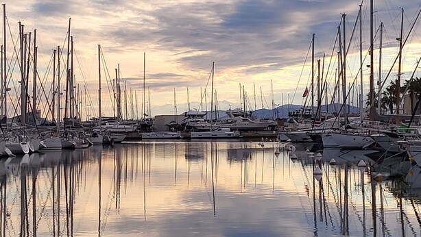 A marina with numerous boats docked, calm water reflecting the boats and sky, and a clear view of the horizon.