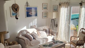 A cozy living room with a sofa, a coffee table, and wicker chairs. There are framed pictures on the wall and a window with curtains.