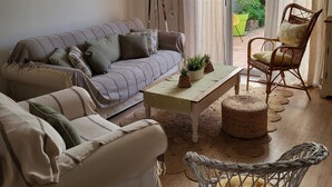 A cozy living room with a sofa, armchair, coffee table, and wicker chair. There are framed pictures on the wall, a lamp, and a view through the curtains.