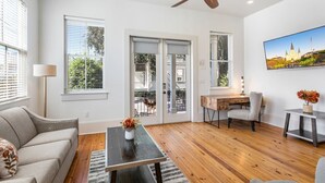 Un salon moderne avec un ventilateur de plafond, une table basse en bois sur laquelle est posé un vase, un canapé, un bureau avec une chaise et une grande fenêtre avec des stores.