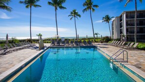 A pool area with clear blue water, surrounded by palm trees and lounge chairs, with a view of the ocean and a building in the background. - Sundial of Sanibel B307, Direct Beachview (Sanibel Island)