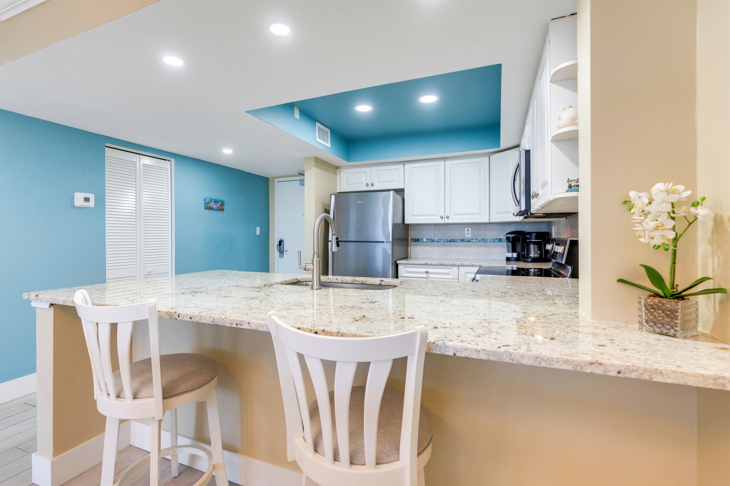 A kitchen with a central island, two white chairs, a refrigerator, and a coffee maker.