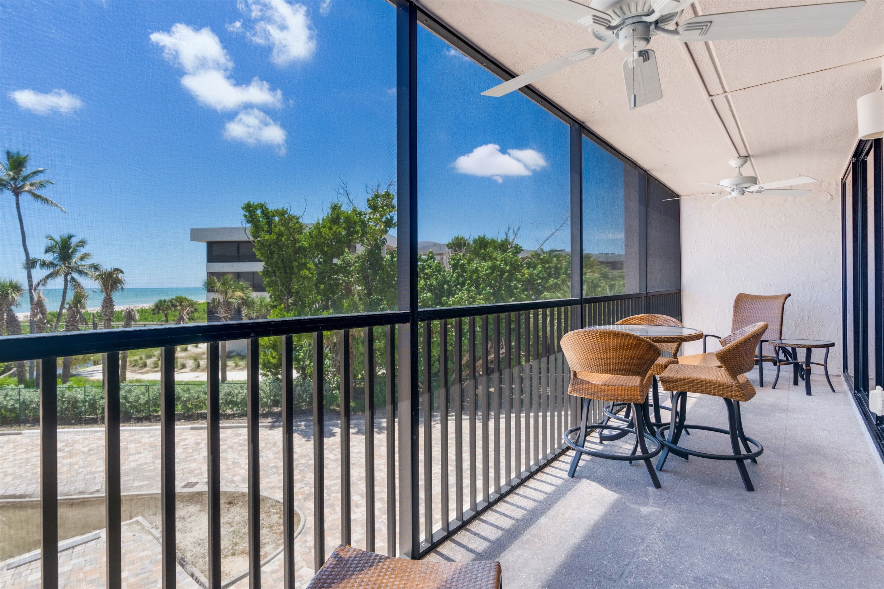A balcony with wicker chairs, a table, and a view of the beach and palm trees.