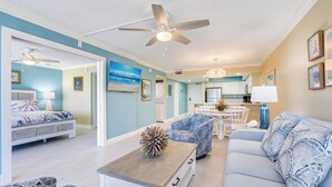 A modern living room with a sofa, a coffee table, and a dining area with a table and chairs. There is a kitchen in the background with cabinets and a refrigerator. - Sundial of Sanibel B307, Direct Beachview (Sanibel Island)