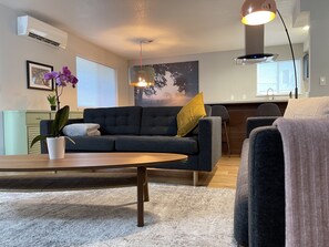 A modern living room with a dark grey sofa, a wooden coffee table, a potted orchid, and a large window with blinds.