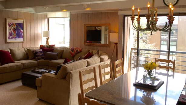 A living room with a sectional sofa, a dining table with chairs, a chandelier, a clock, and a window with a view of trees. - Pristine Condo, Walk To Lifts, 2 Fireplaces, 3 Balconies,  Mountain Views! (Vail)