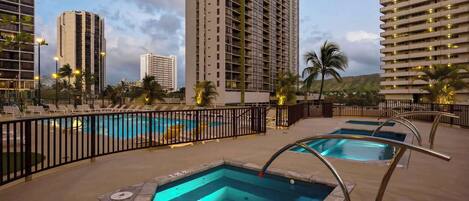 A hotel pool area with multiple pools, a black metal fence, and tall buildings in the background.