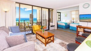 A hotel room with a balcony, a sofa, a coffee table, a TV, and a bed with blue bedding. - Waikiki Banyan condo - ocean views & beach access (Honolulu)