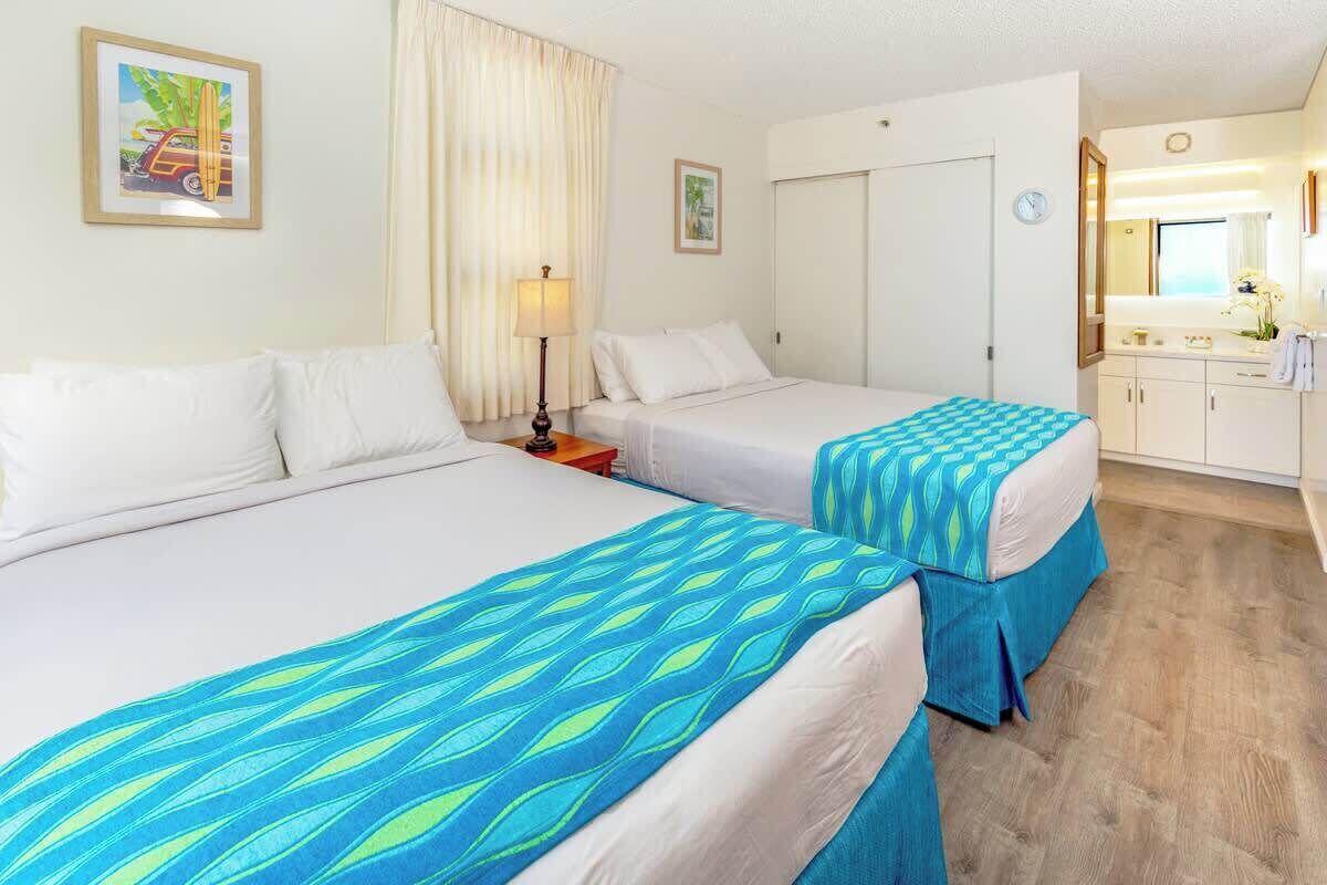 A hotel room with two beds, a desk, a chair, a lamp, a clock, and a bathroom with a mirror and sink.
